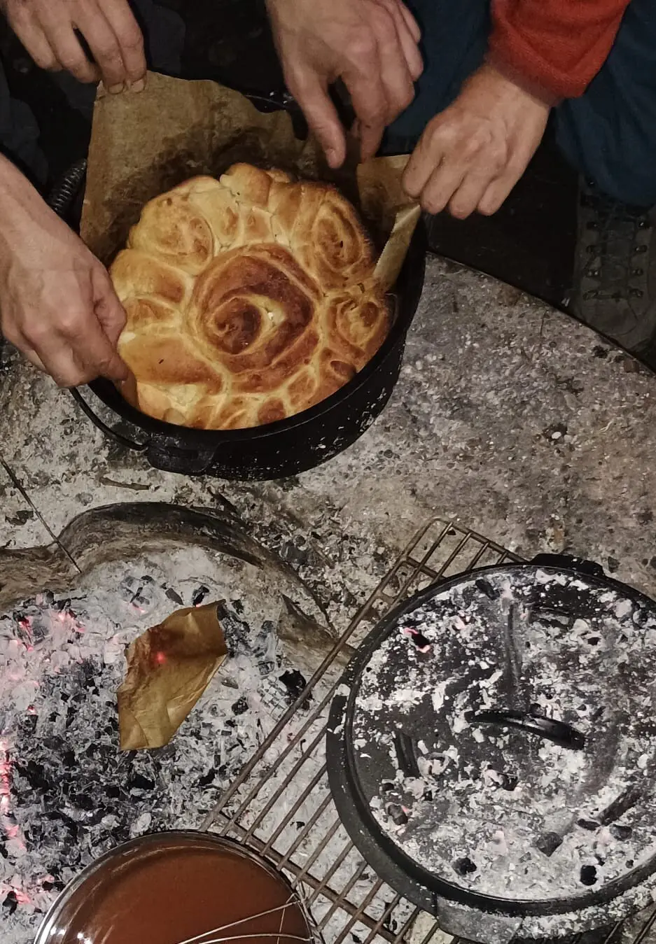 Mehrere Hände nehmen ein rundes, goldbraun gebackenes Brot aus einem schwarzen Topf über einer Feuerstelle mit glühender Asche | © DAV-FN / M. Gross