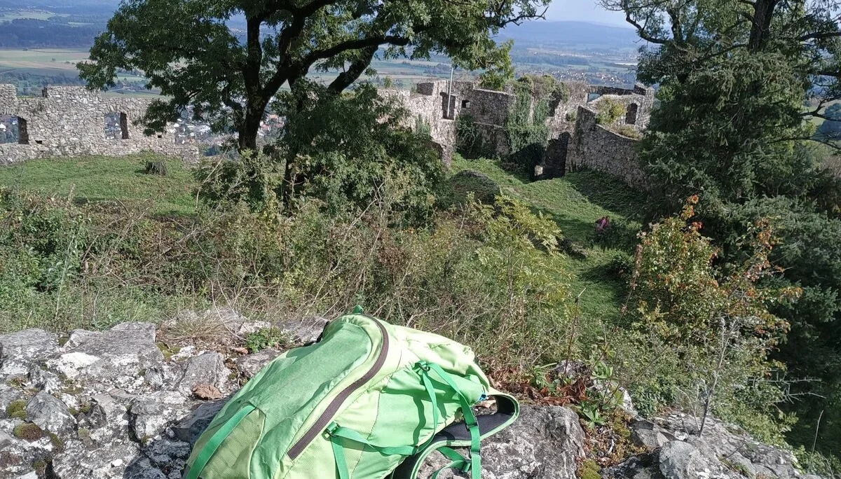 Grüner Rucksack liegt auf Felsen, im Hintergrund Ruinen einer Burg und Bäume unter blauem Himmel | © DAV-FN / M. Gross