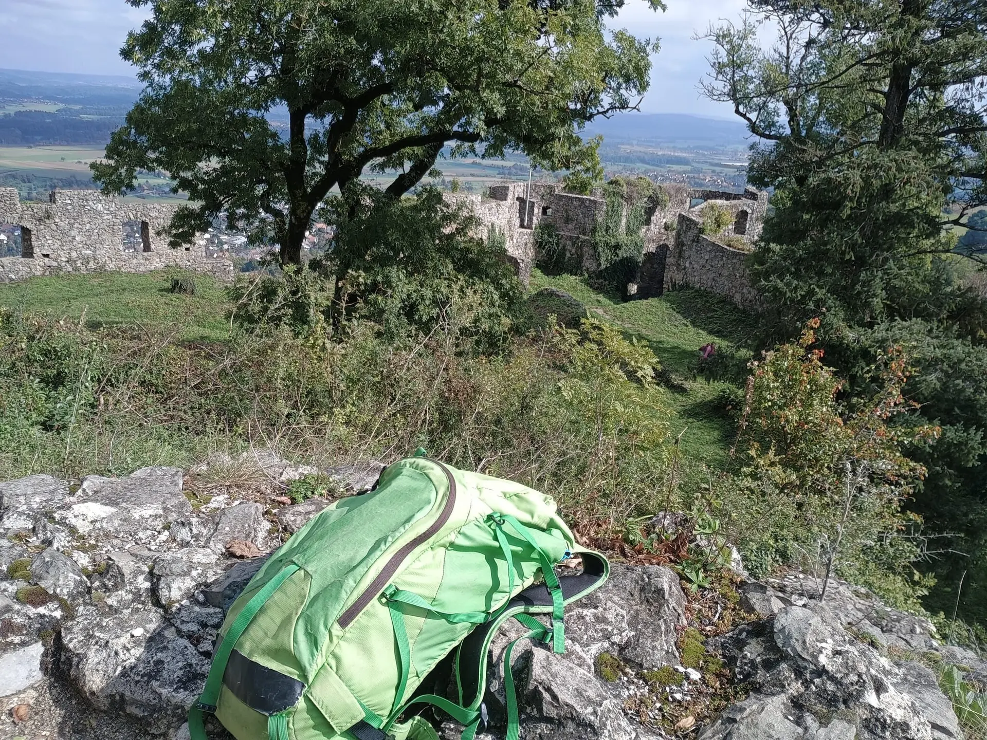 Grüner Rucksack liegt auf Felsen, im Hintergrund Ruinen einer Burg und Bäume unter blauem Himmel | © DAV-FN / M. Gross