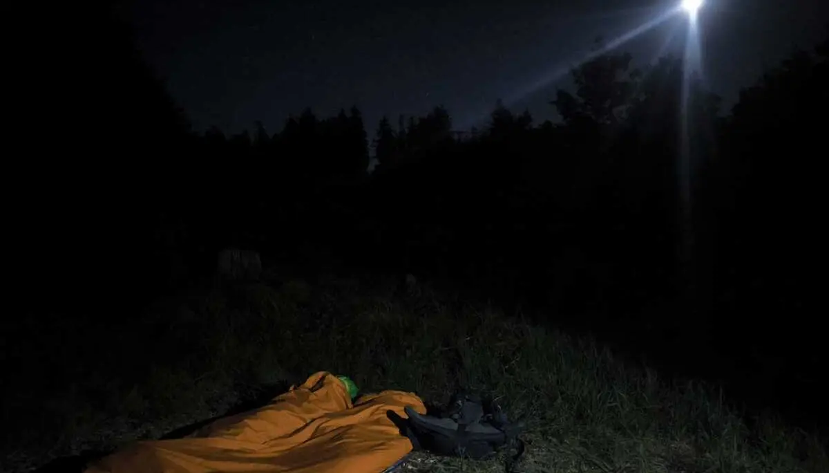 Person liegt in einem orangenen Schlafsack auf einer Wiese unter einem dunklen Nachthimmel mit hellem Mond | © DAV-FN / M. Gross