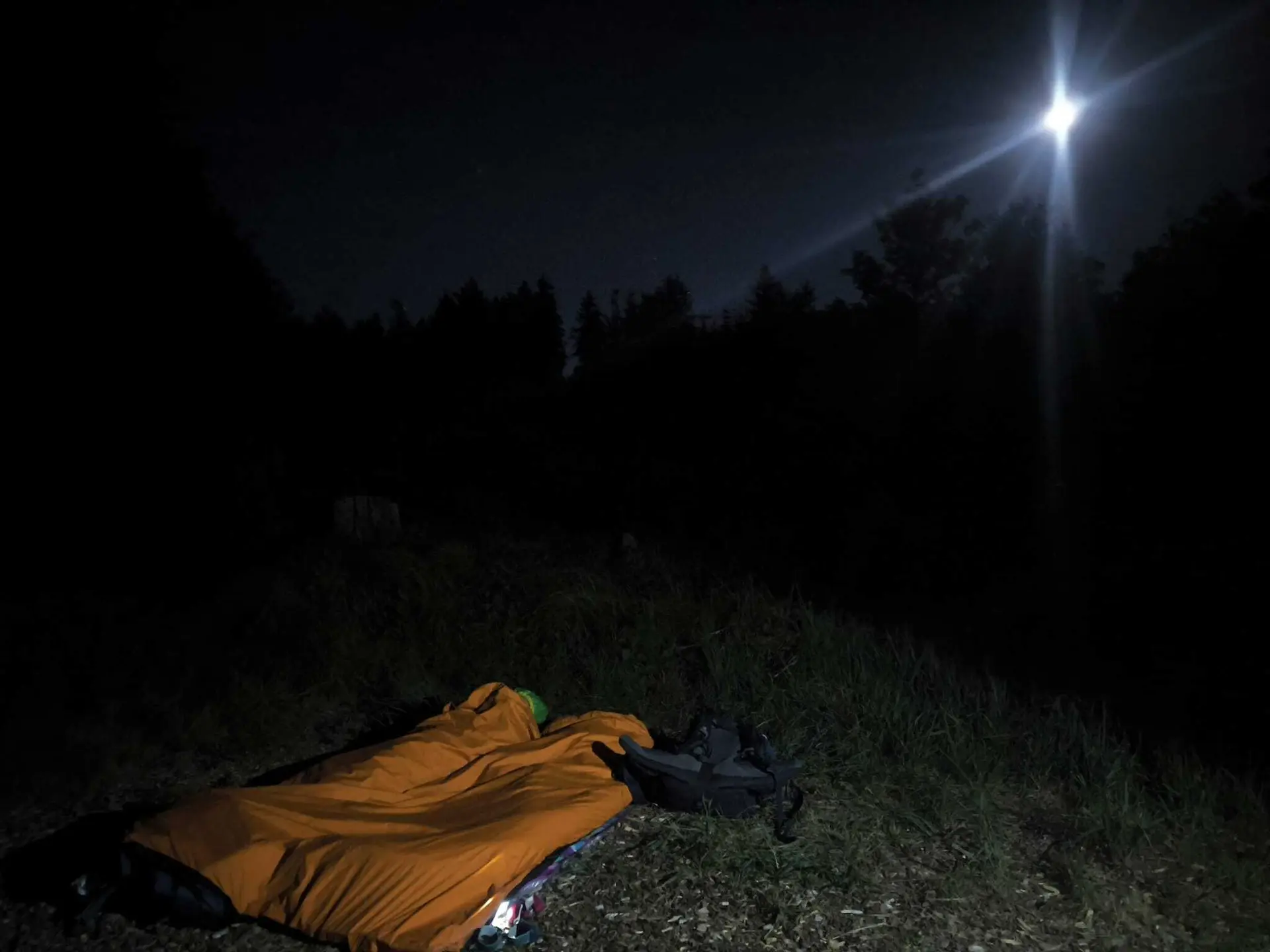 Person liegt in einem orangenen Schlafsack auf einer Wiese unter einem dunklen Nachthimmel mit hellem Mond | © DAV-FN / M. Gross
