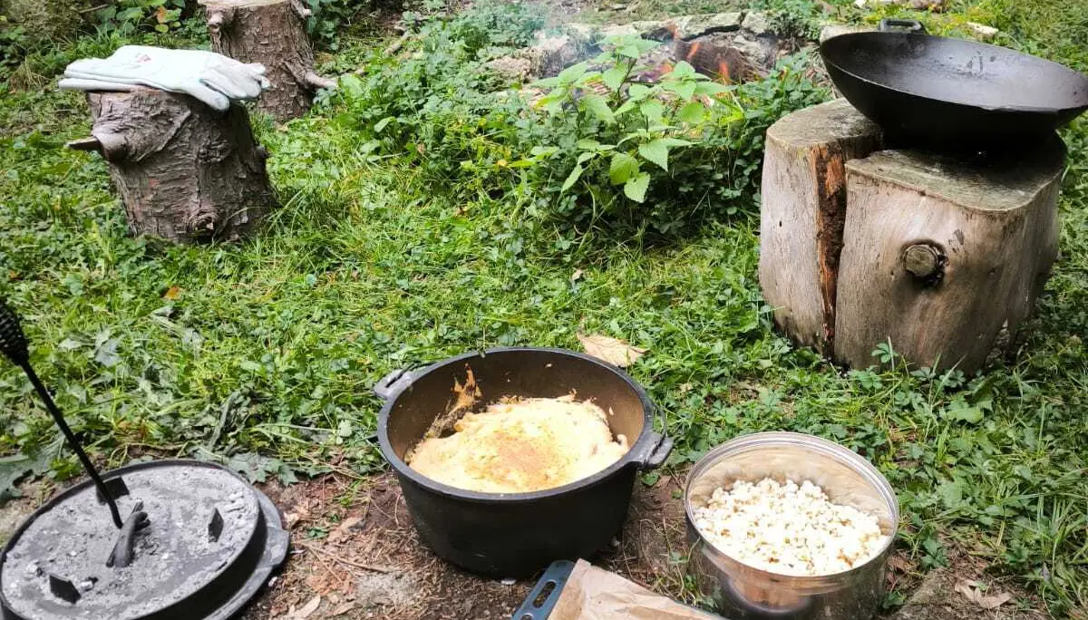 Grillstelle im Grünen mit rauchendem Feuer, Gusseisentopf, Schale mit Popcorn und Backblech mit Nüssen auf dem Boden | © DAV-FN / M.Groß