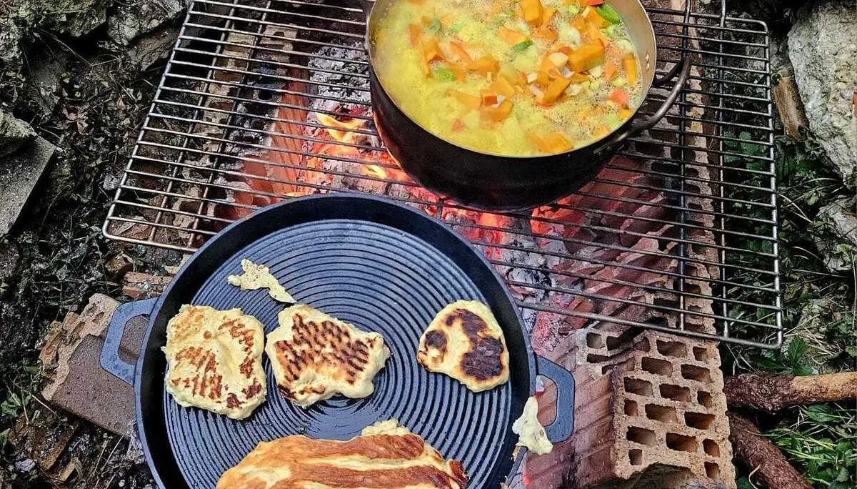 Topf mit Gemüsesuppe und Pfanne mit verschiedenen Teigfladen auf Grillrost über offenem Feuer im Freien | © DAV-FN / M.Groß