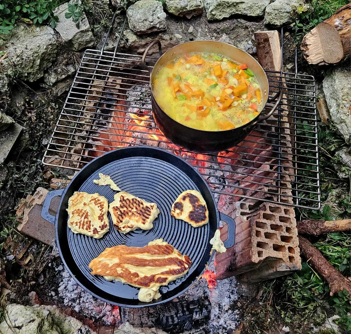 Topf mit Gemüsesuppe und Pfanne mit verschiedenen Teigfladen auf Grillrost über offenem Feuer im Freien | © DAV-FN / M.Groß
