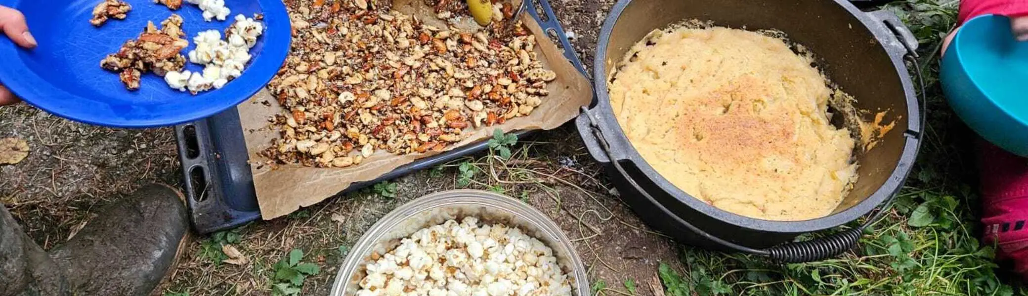 Zwei Personen bereiten im Freien auf einer Unterlage Nüsse und Popcorn zu, daneben steht ein Topf mit Maisbrei | © DAV-FN / M.Groß