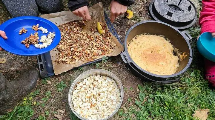 Zwei Personen bereiten im Freien auf einer Unterlage Nüsse und Popcorn zu, daneben steht ein Topf mit Maisbrei | © DAV-FN / M.Groß