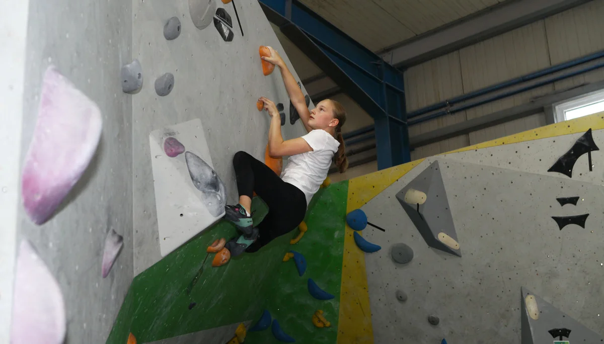 Jugendliche klettert an einer Indoor-Kletterwand mit bunten Griffen, trägt weiße T-Shirt und schwarze Hose | © DAV-FN Thomas Müller