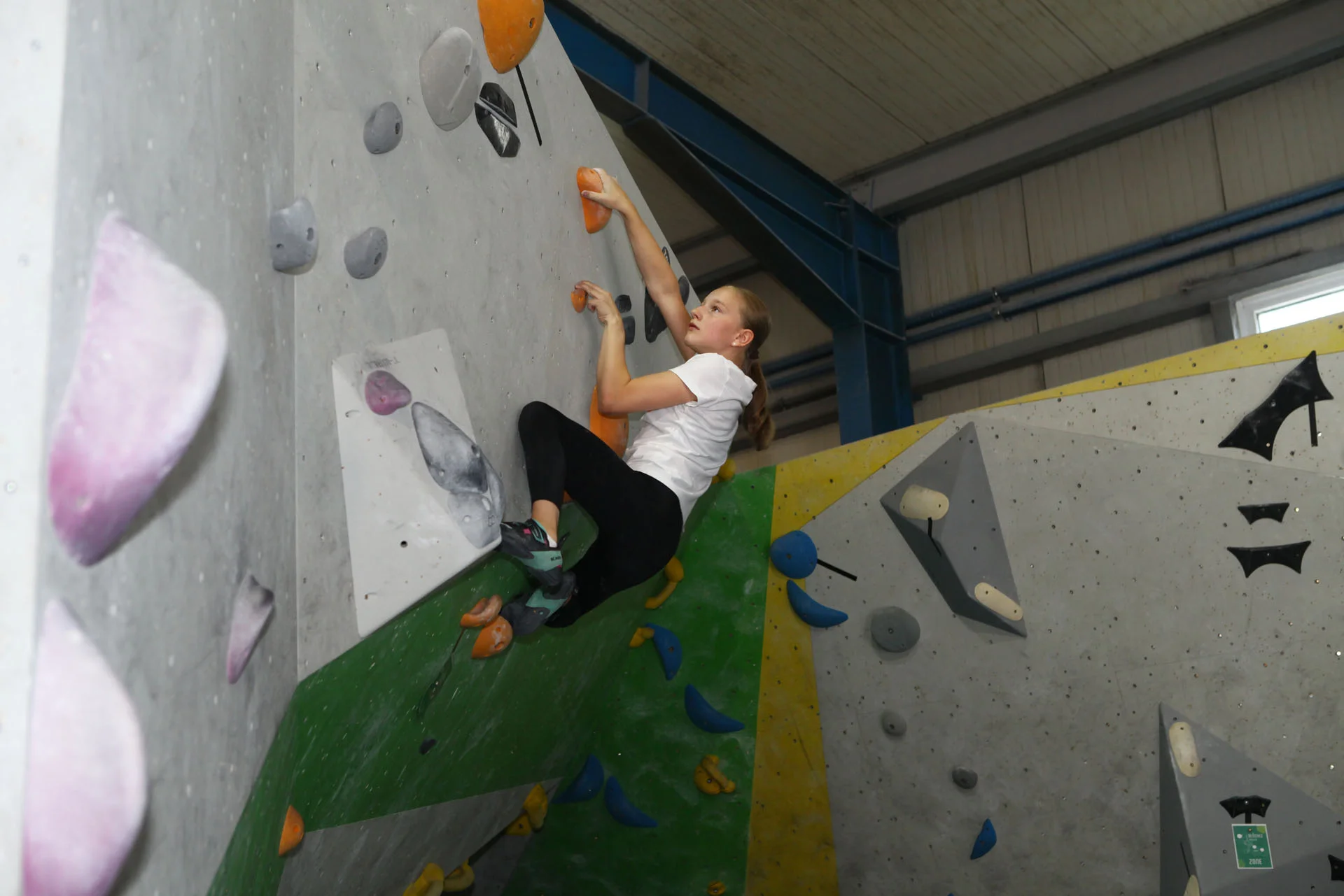 Jugendliche klettert an einer Indoor-Kletterwand mit bunten Griffen, trägt weiße T-Shirt und schwarze Hose | © DAV-FN Thomas Müller