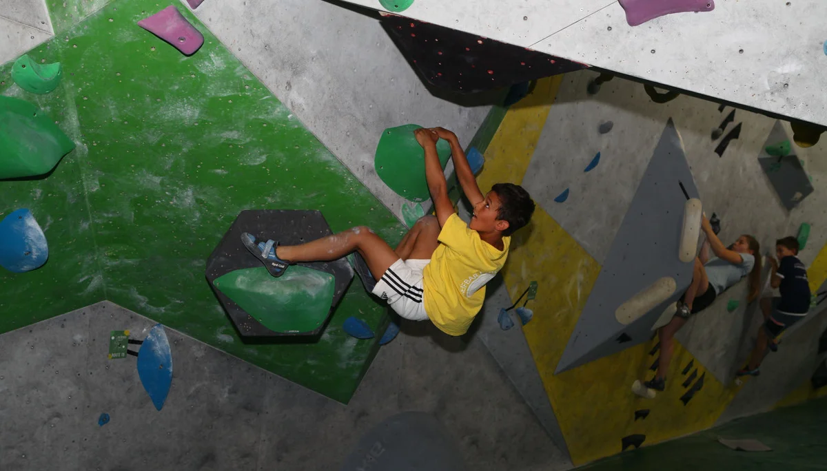 Kind in gelbem T-Shirt klettert an einer Indoor-Boulderwand mit grünen und schwarzen Griffen | © DAV-FN Thomas Müller