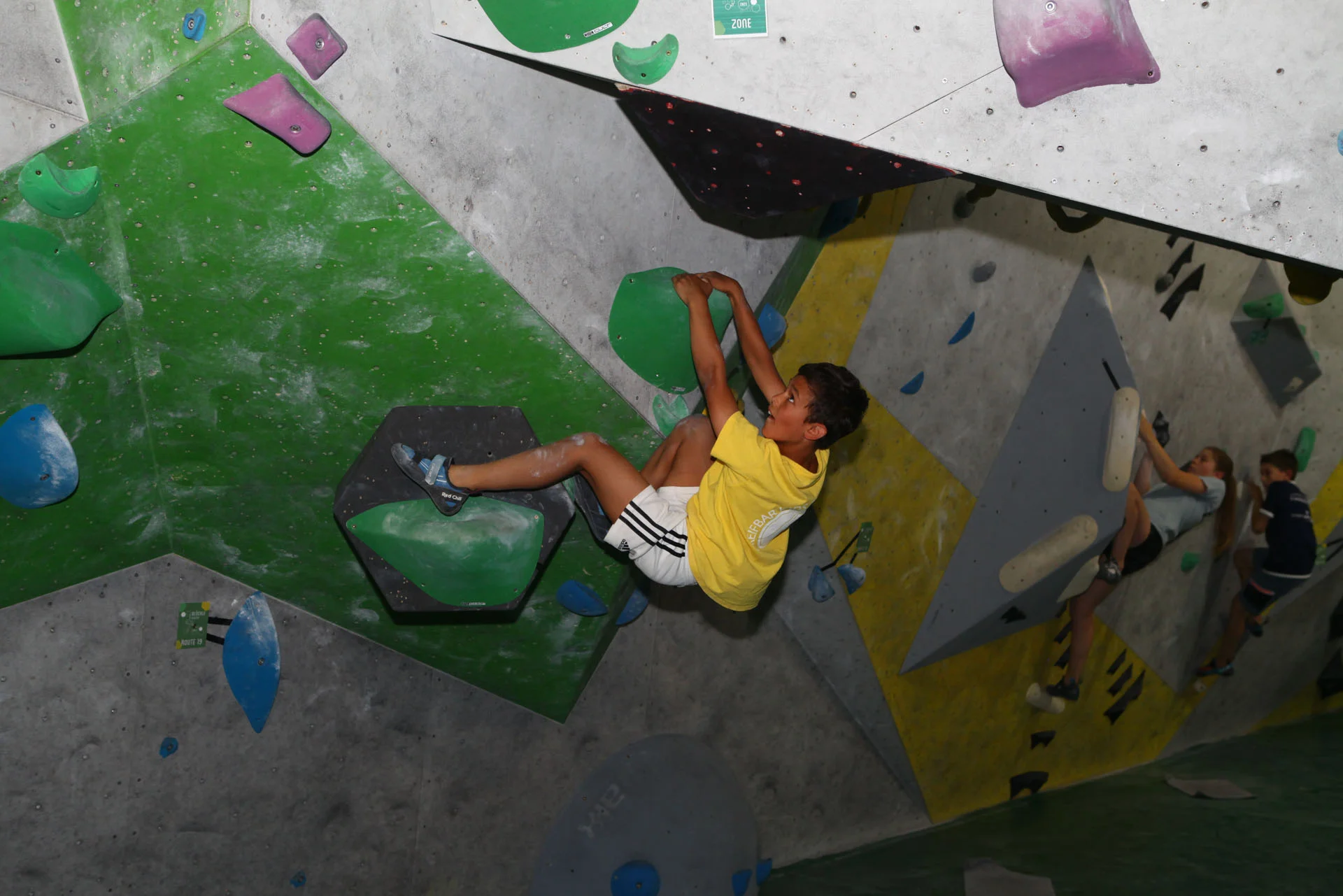 Kind in gelbem T-Shirt klettert an einer Indoor-Boulderwand mit grünen und schwarzen Griffen | © DAV-FN Thomas Müller