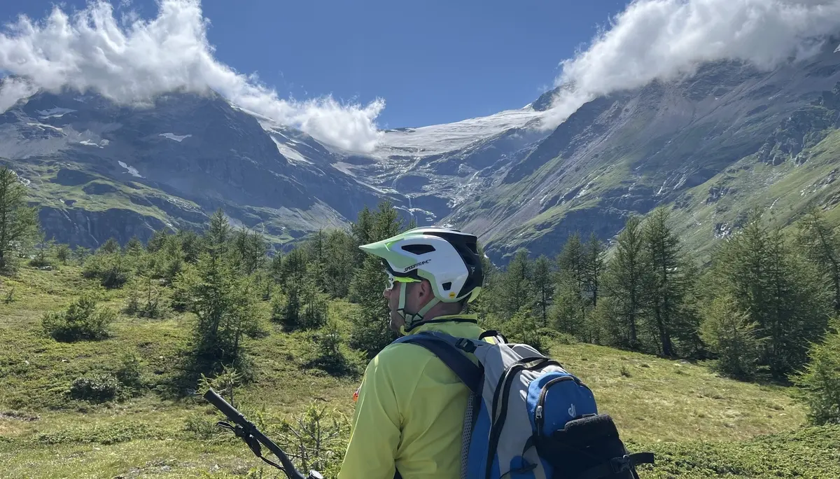Person mit Fahrradhelm und Rucksack steht auf einer Wiese vor bewaldeten Bergen unter blauem Himmel mit Wolken | © DAV-FN / E. Flöth, M. Arzner, S. Haberer 