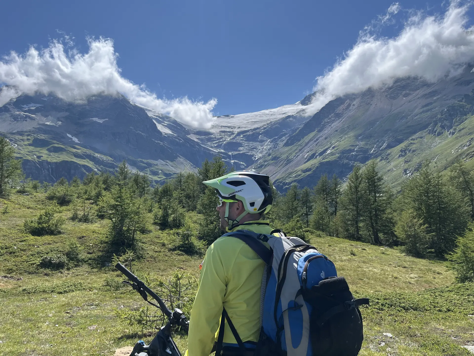 Person mit Fahrradhelm und Rucksack steht auf einer Wiese vor bewaldeten Bergen unter blauem Himmel mit Wolken | © DAV-FN / E. Flöth, M. Arzner, S. Haberer 