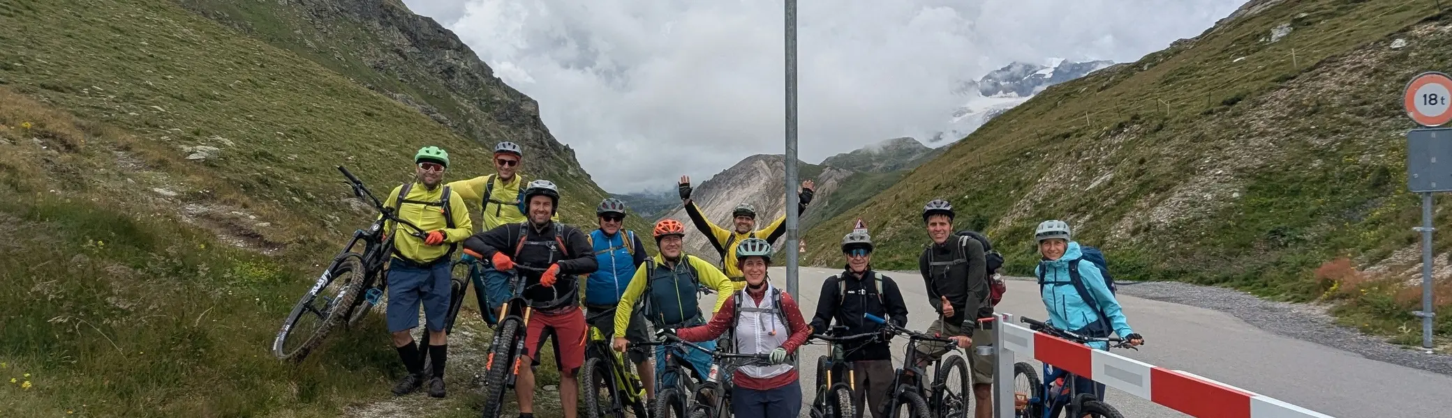 Gruppe von Mountainbikern mit Helmen und Fahrrädern steht an einem Passschild auf einer Bergstraße zwischen grünen Hängen | © DAV-FN / E. Flöth, M. Arzner, S. Haberer 