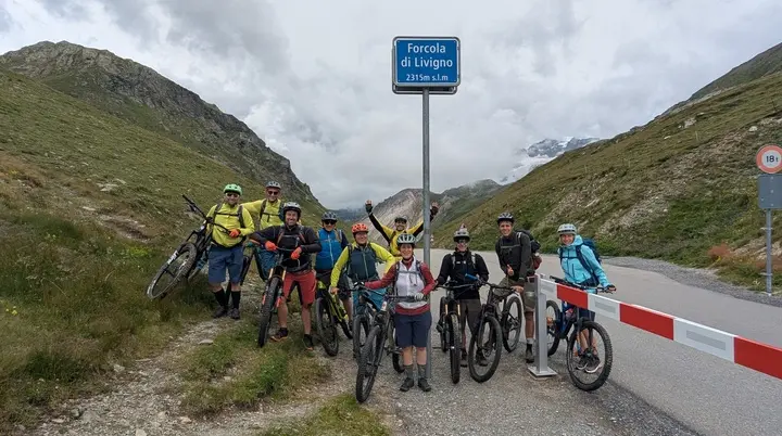 Gruppe von Mountainbikern mit Helmen und Fahrrädern steht an einem Passschild auf einer Bergstraße zwischen grünen Hängen | © DAV-FN / E. Flöth, M. Arzner, S. Haberer 