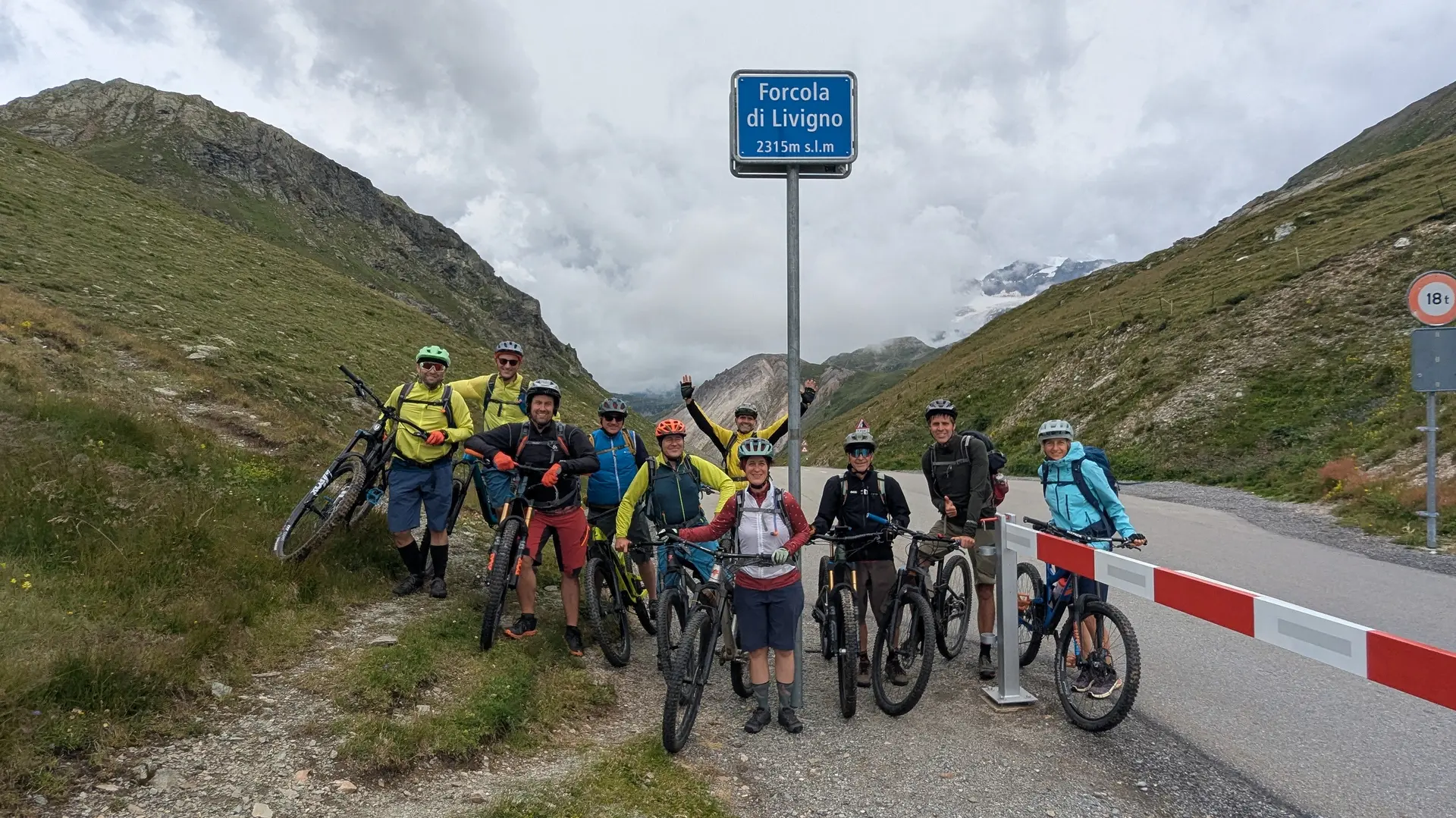 Gruppe von Mountainbikern mit Helmen und Fahrrädern steht an einem Passschild auf einer Bergstraße zwischen grünen Hängen | © DAV-FN / E. Flöth, M. Arzner, S. Haberer 