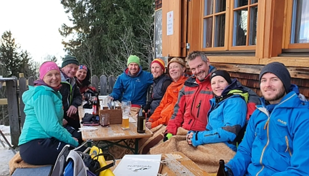 Mehrere Personen in bunter Outdoor-Bekleidung sitzen an einem Holztisch vor einer Hütte mit geöffneten Fensterläden. | © DAV-FN / Peter Schäfer