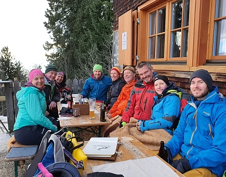 Mehrere Personen in bunter Outdoor-Bekleidung sitzen an einem Holztisch vor einer Hütte mit geöffneten Fensterläden. | © DAV-FN / Peter Schäfer