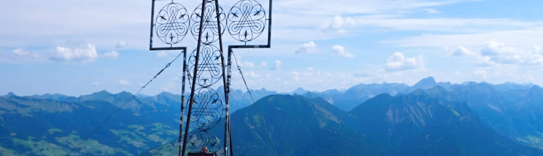 Blick am Gipfelkreuz der Mondspitze vorbei auf die Alpenstadt Budenz und die Berge des Bregenzer Waldes | © Foto Walter Biselli, CC BY-SA,