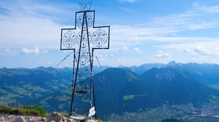 Blick am Gipfelkreuz der Mondspitze vorbei auf die Alpenstadt Budenz und die Berge des Bregenzer Waldes | © Foto Walter Biselli, CC BY-SA,