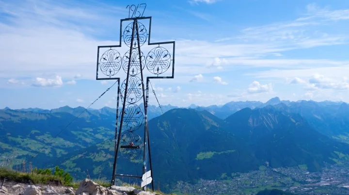 Blick am Gipfelkreuz der Mondspitze vorbei auf die Alpenstadt Budenz und die Berge des Bregenzer Waldes | © Foto Walter Biselli, CC BY-SA,