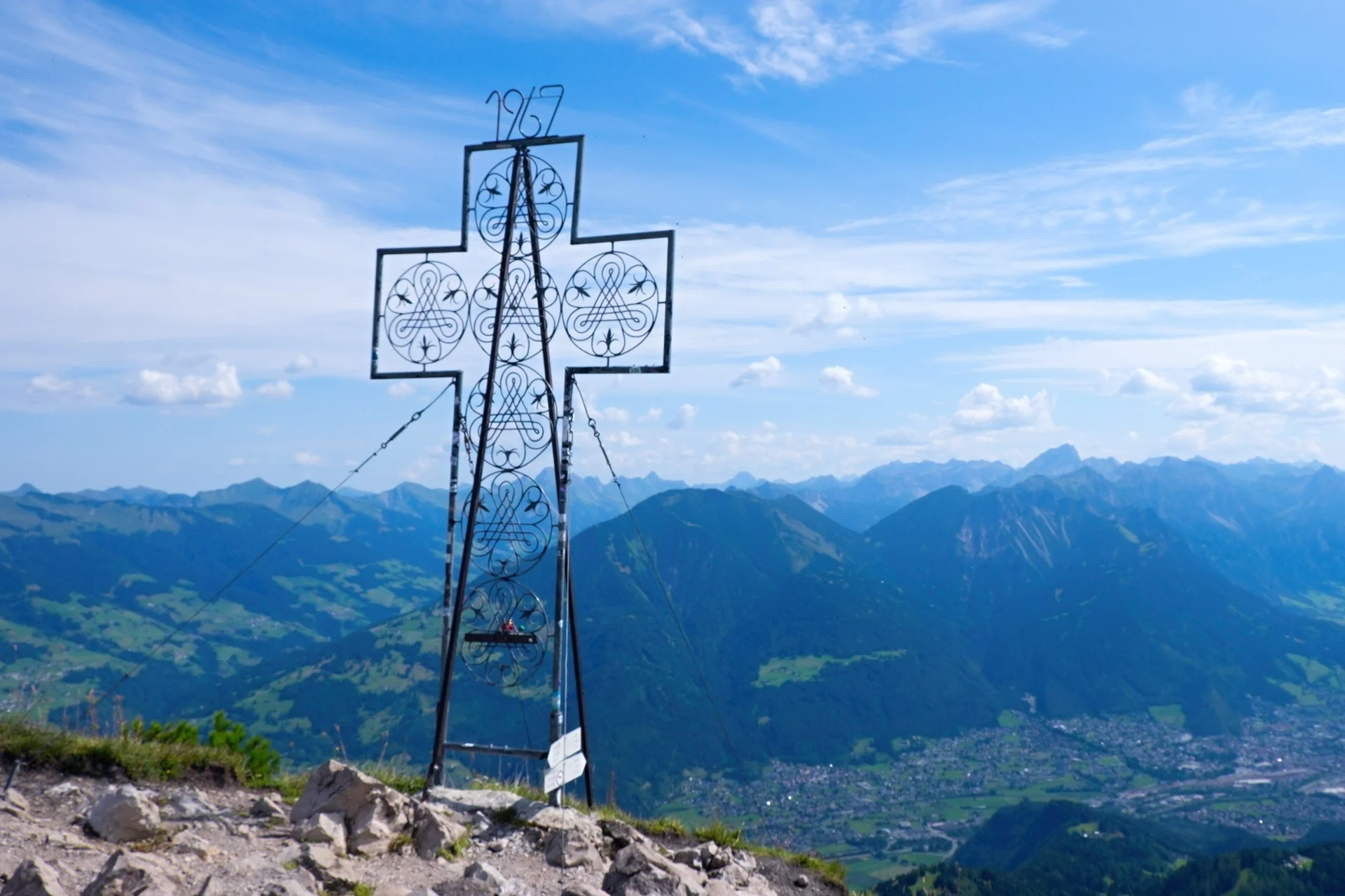 Blick am Gipfelkreuz der Mondspitze vorbei auf die Alpenstadt Budenz und die Berge des Bregenzer Waldes | © Foto Walter Biselli, CC BY-SA,