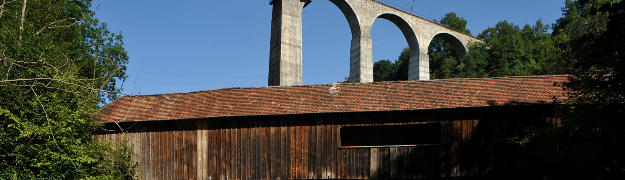 Holzbrücke mit Ziegeldach vor einer hohen Steinbogenbrücke mit Eisenbahnstrecke in bewaldeter Landschaft | © Хрюша - Eigenes Werk, CC BY-SA 3.0, httpscommons.wikimedia.orgwindex.phpcurid=7596023