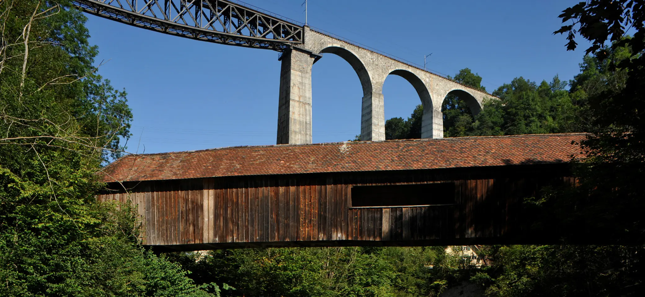 Holzbrücke mit Ziegeldach vor einer hohen Steinbogenbrücke mit Eisenbahnstrecke in bewaldeter Landschaft | © Хрюша - Eigenes Werk, CC BY-SA 3.0, httpscommons.wikimedia.orgwindex.phpcurid=7596023