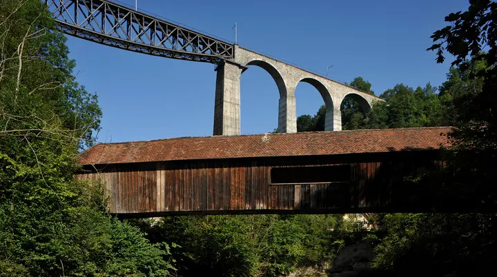 Holzbrücke mit Ziegeldach vor einer hohen Steinbogenbrücke mit Eisenbahnstrecke in bewaldeter Landschaft | © Хрюша - Eigenes Werk, CC BY-SA 3.0, httpscommons.wikimedia.orgwindex.phpcurid=7596023