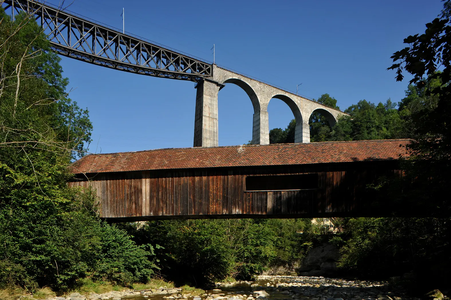 Holzbrücke mit Ziegeldach vor einer hohen Steinbogenbrücke mit Eisenbahnstrecke in bewaldeter Landschaft | © Хрюша - Eigenes Werk, CC BY-SA 3.0, httpscommons.wikimedia.orgwindex.phpcurid=7596023