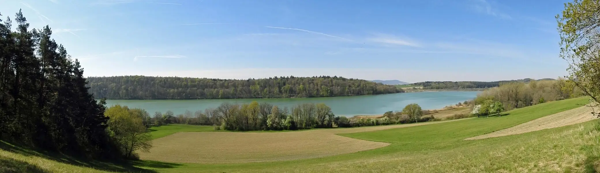 Panoramaaufnahme eines Sees mit bewaldetem Ufer und Wiesen im Vordergrund unter blauem Himmel | © Von Flodur63  CC BY-SA 4.0, Wikipedia
