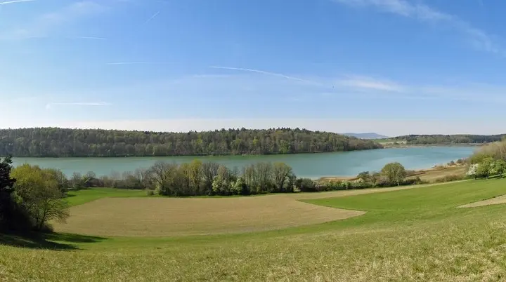 Panoramaaufnahme eines Sees mit bewaldetem Ufer und Wiesen im Vordergrund unter blauem Himmel | © Von Flodur63  CC BY-SA 4.0, Wikipedia
