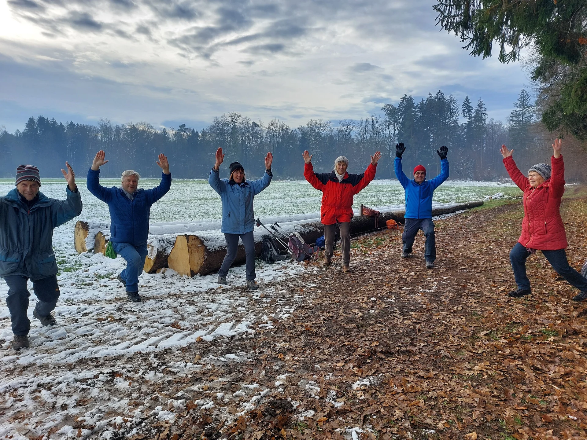 Gruppe von sieben Personen in Winterkleidung, die auf einem schneebedeckten Feld und einem Waldweg stehen und die Arme heben. | © DAV-FN / G. Wocher
