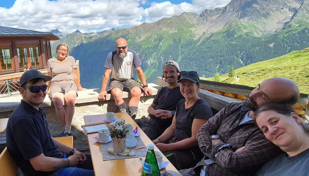 Sieben Personen sitzen an einem Holztisch auf der Terrasse der Häfler Hütte mit Bergpanorama und blauem Himmel mit Wolken | © DAV-FN / R. Klees o. R. Zahner