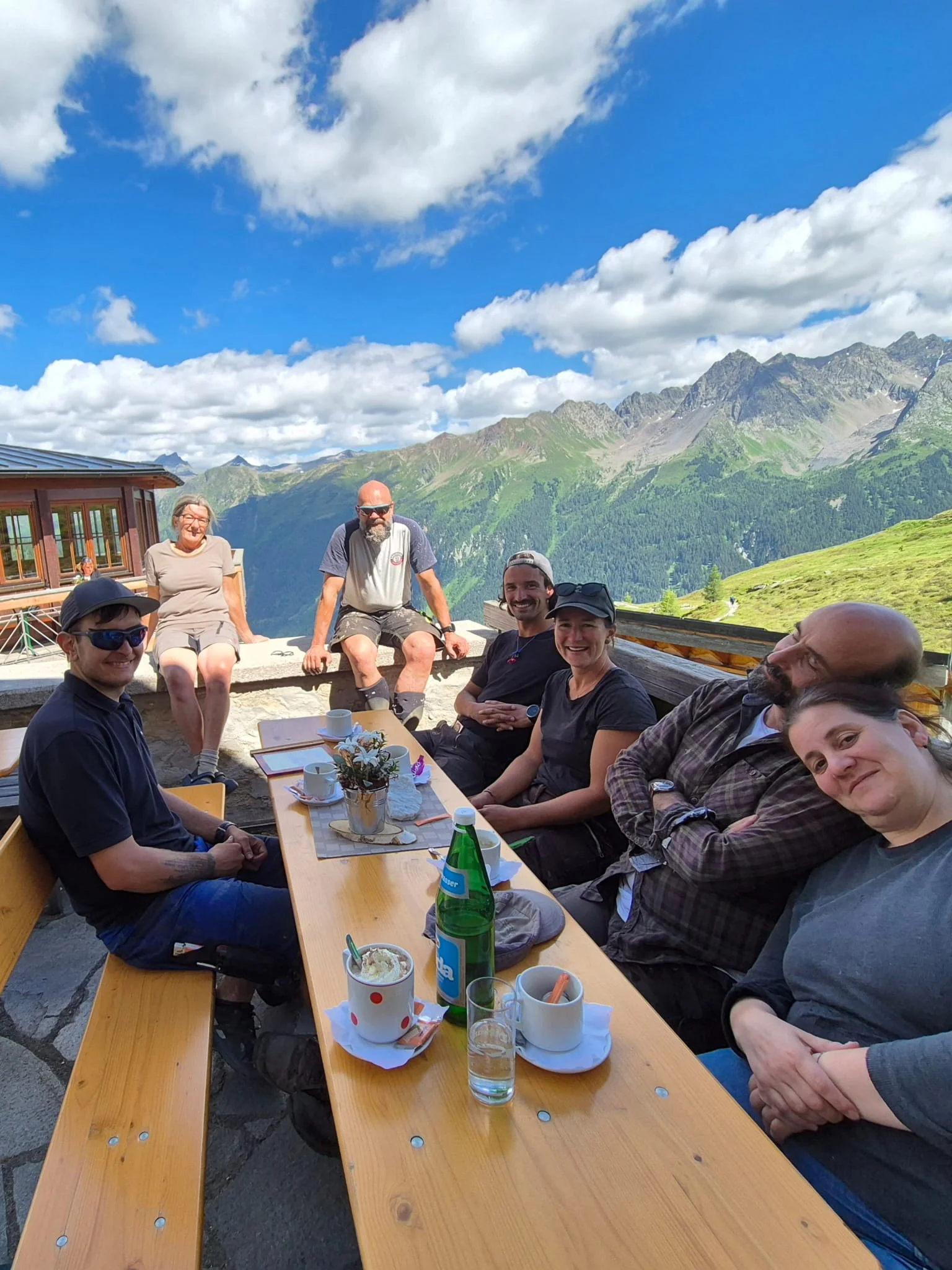 Sieben Personen sitzen an einem Holztisch auf der Terrasse der Häfler Hütte mit Bergpanorama und blauem Himmel mit Wolken | © DAV-FN / R. Klees o. R. Zahner
