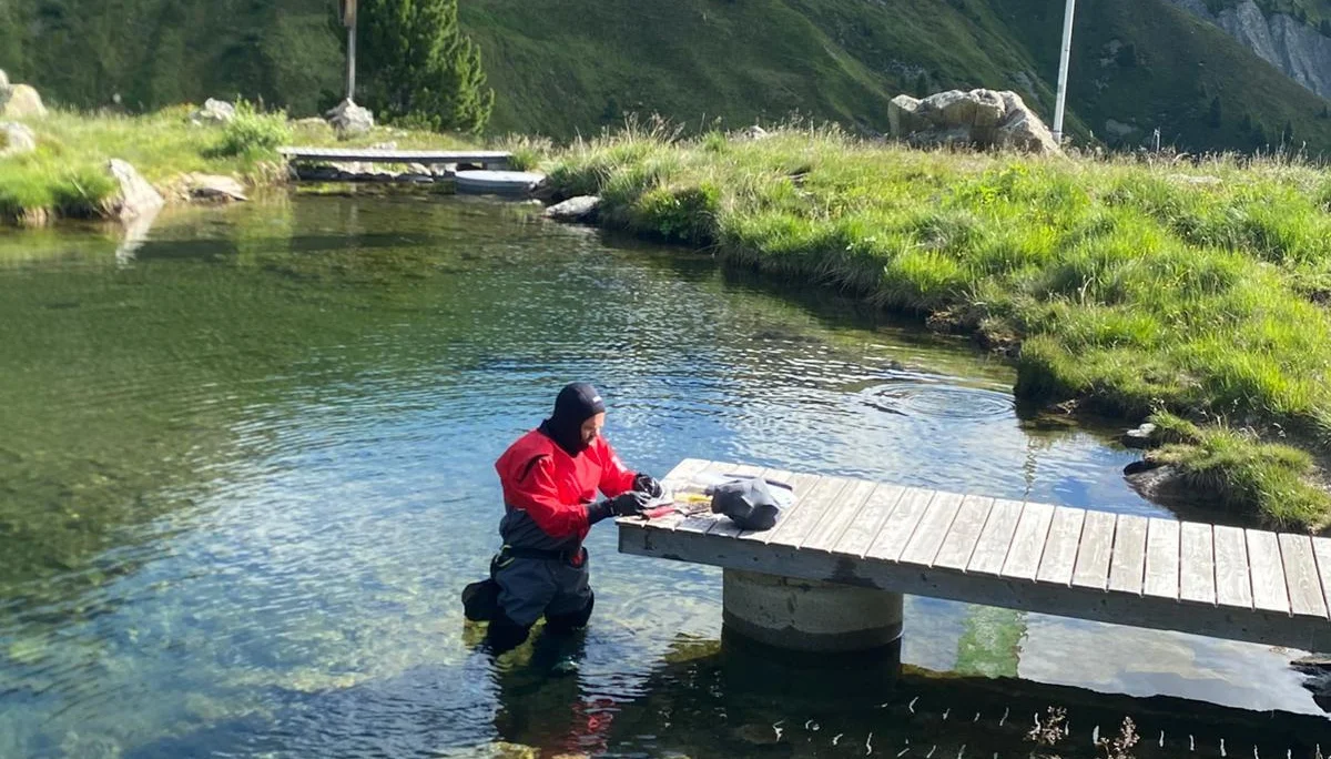 Person in Tauchanzug steht im klaren Wasser eines Bergsees neben einem Holzsteg, umgeben von grünen Bergen und einer Fahne mit Text | © DAV-FN / R. Klees o. R. Zahner