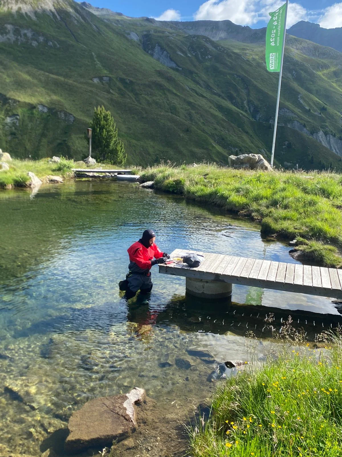 Person in Tauchanzug steht im klaren Wasser eines Bergsees neben einem Holzsteg, umgeben von grünen Bergen und einer Fahne mit Text | © DAV-FN / R. Klees o. R. Zahner