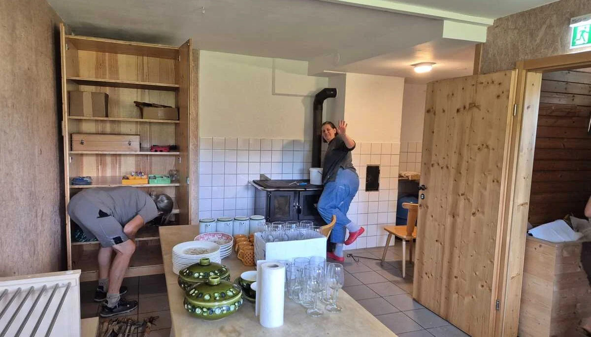 Zwei Personen in einem rustikalen Winterraum mit Holzofen, Regalen und einem Tisch mit Geschirr und Gläsern | © DAV-FN / R. Klees o. R. Zahner