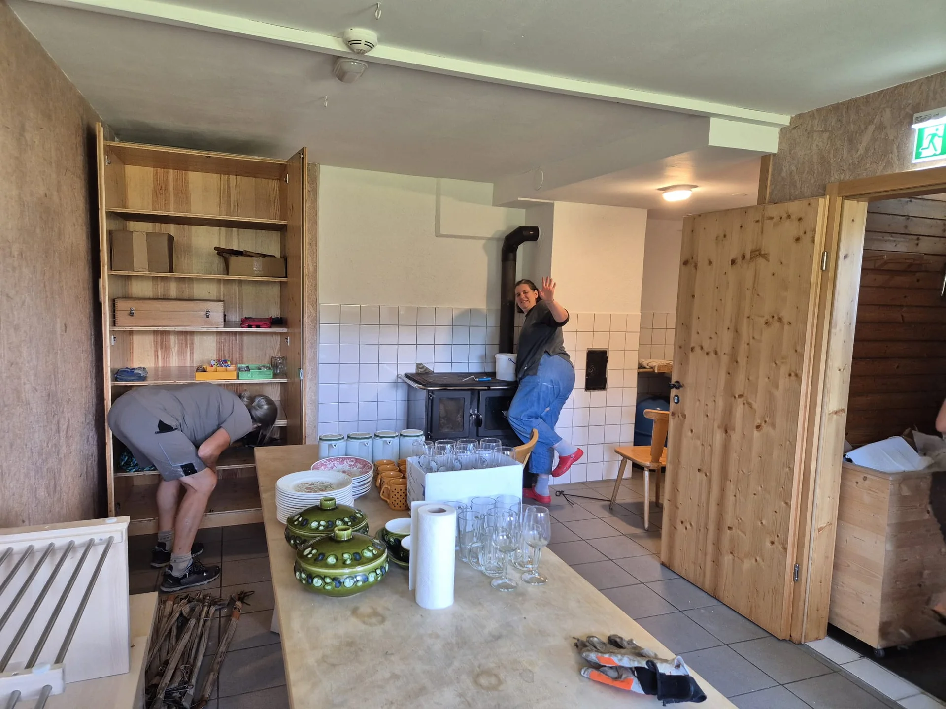 Zwei Personen in einem rustikalen Winterraum mit Holzofen, Regalen und einem Tisch mit Geschirr und Gläsern | © DAV-FN / R. Klees o. R. Zahner