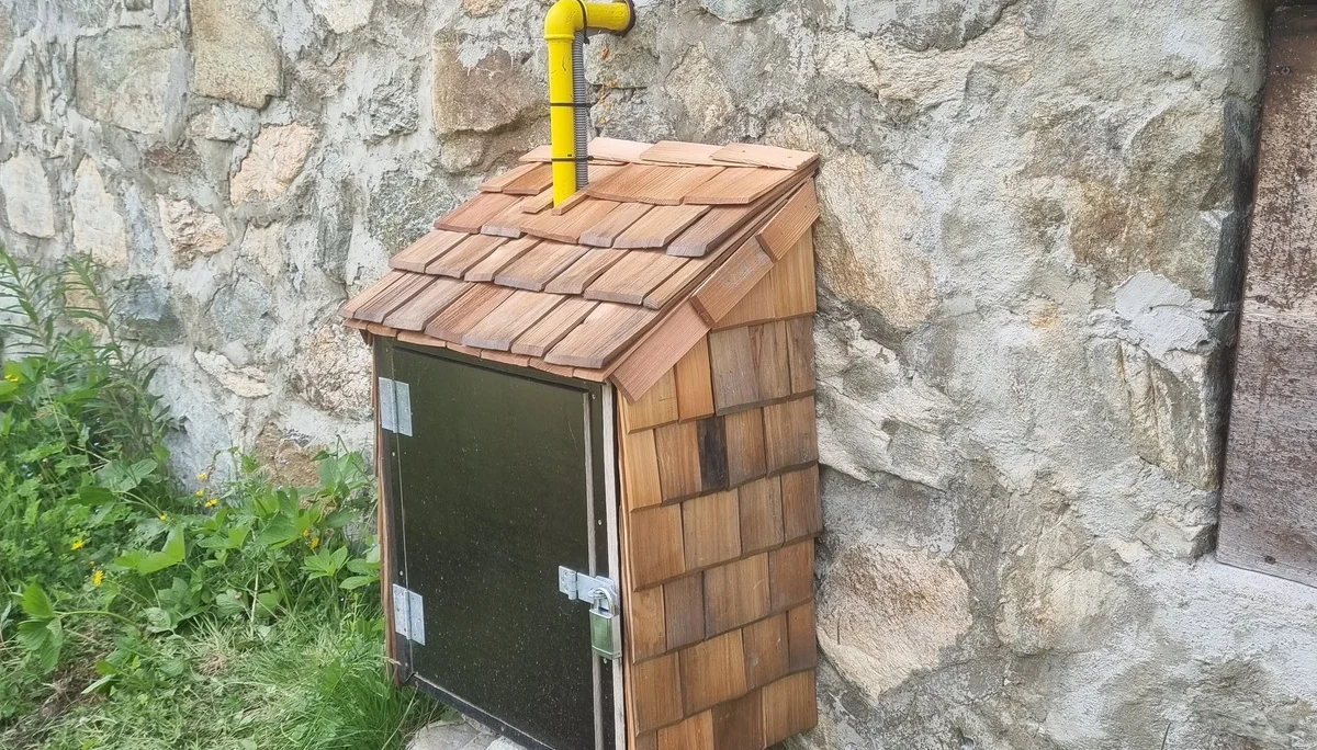 Kleines Holzhaus mit Schindeldach und gelbem Rohr, befestigt an einer Steinmauer, mit Metalltür und Steinen als Fundament | © DAV-FN / R. Klees o. R. Zahner