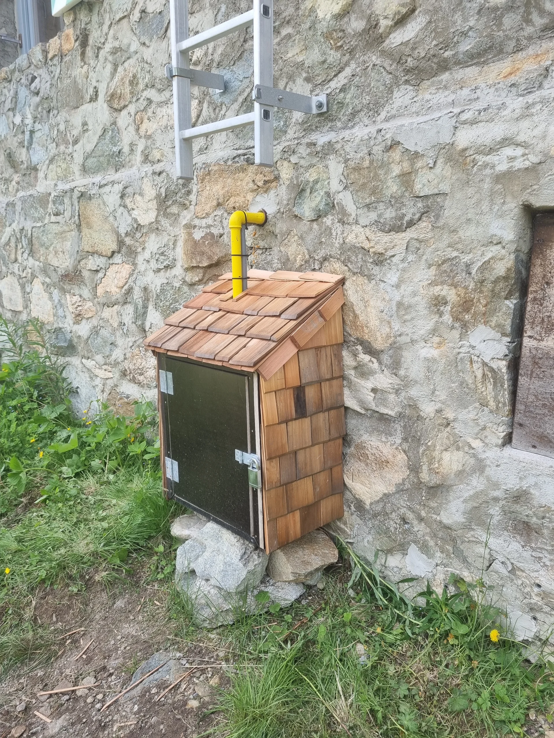 Kleines Holzhaus mit Schindeldach und gelbem Rohr, befestigt an einer Steinmauer, mit Metalltür und Steinen als Fundament | © DAV-FN / R. Klees o. R. Zahner