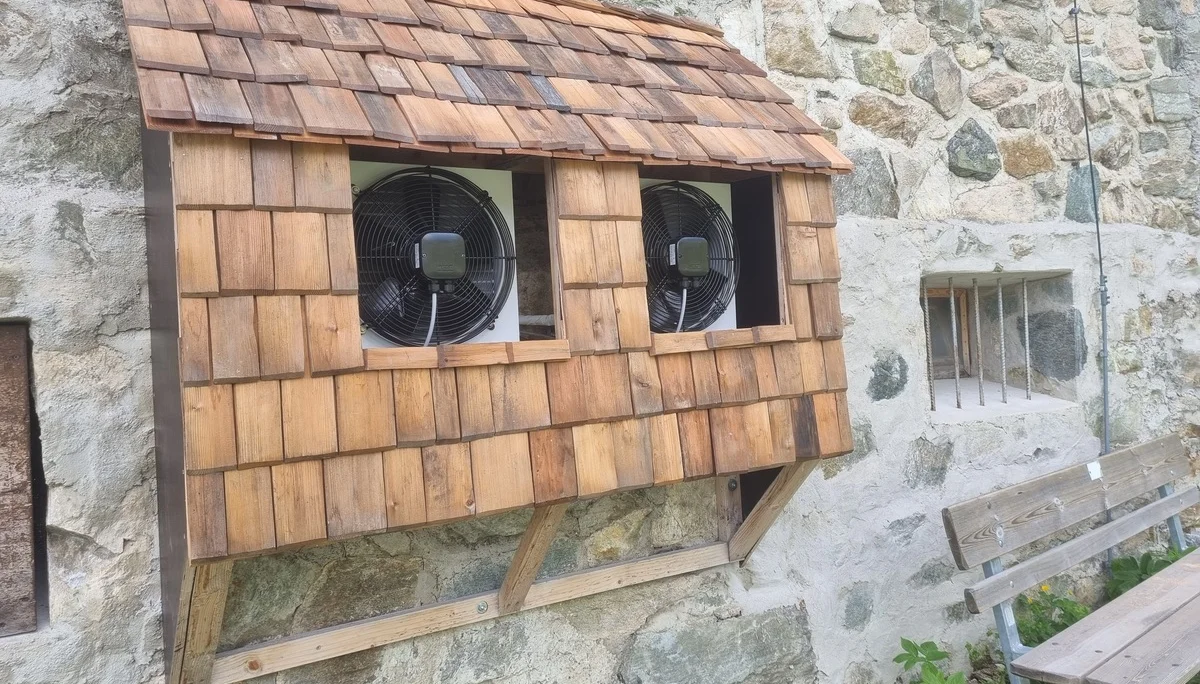 Holzverkleidung mit zwei eingebauten Ventilatoren an einer Steinmauer, daneben eine Holzbank | © DAV-FN / R. Klees o. R. Zahner