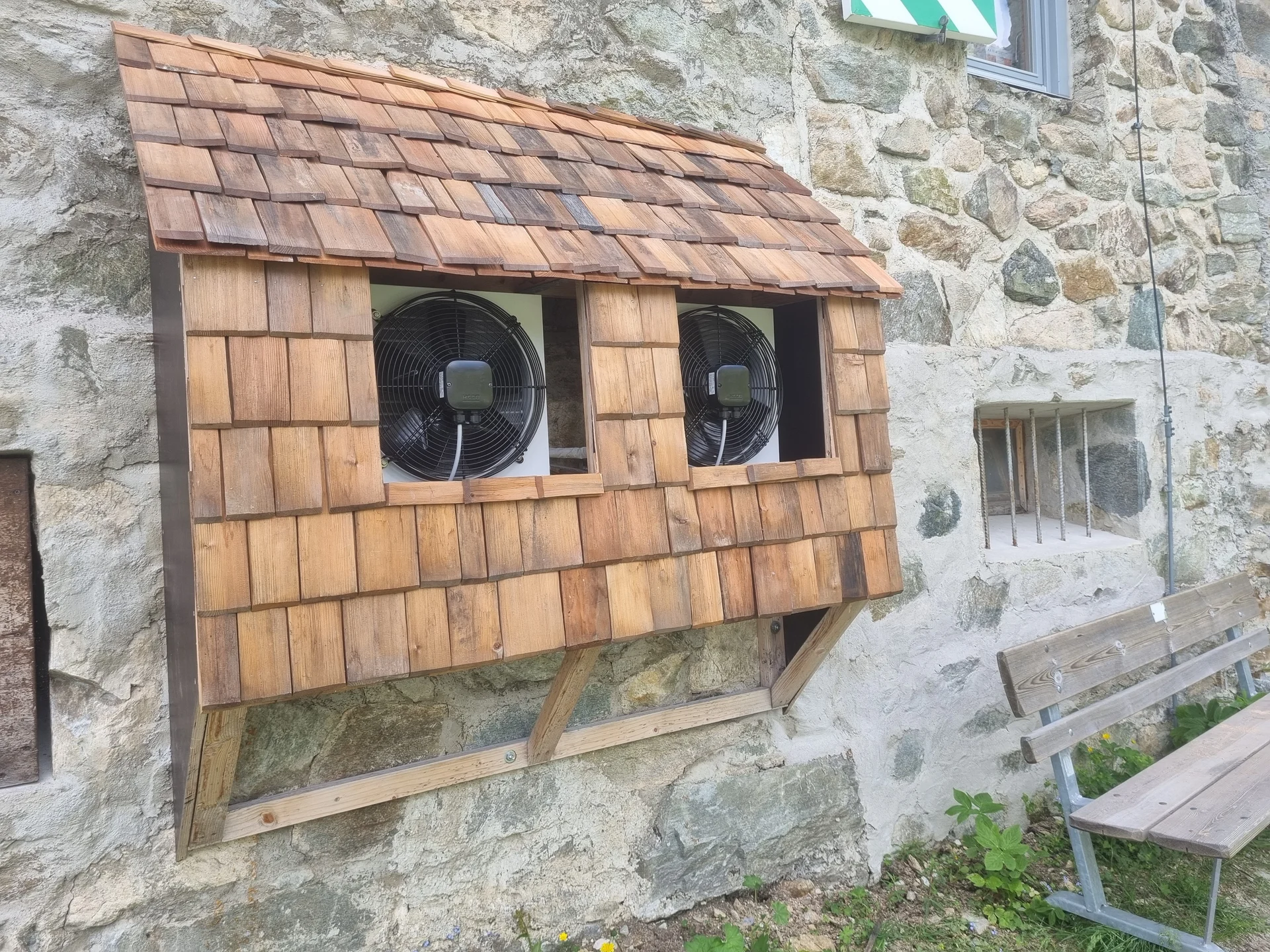 Holzverkleidung mit zwei eingebauten Ventilatoren an einer Steinmauer, daneben eine Holzbank | © DAV-FN / R. Klees o. R. Zahner