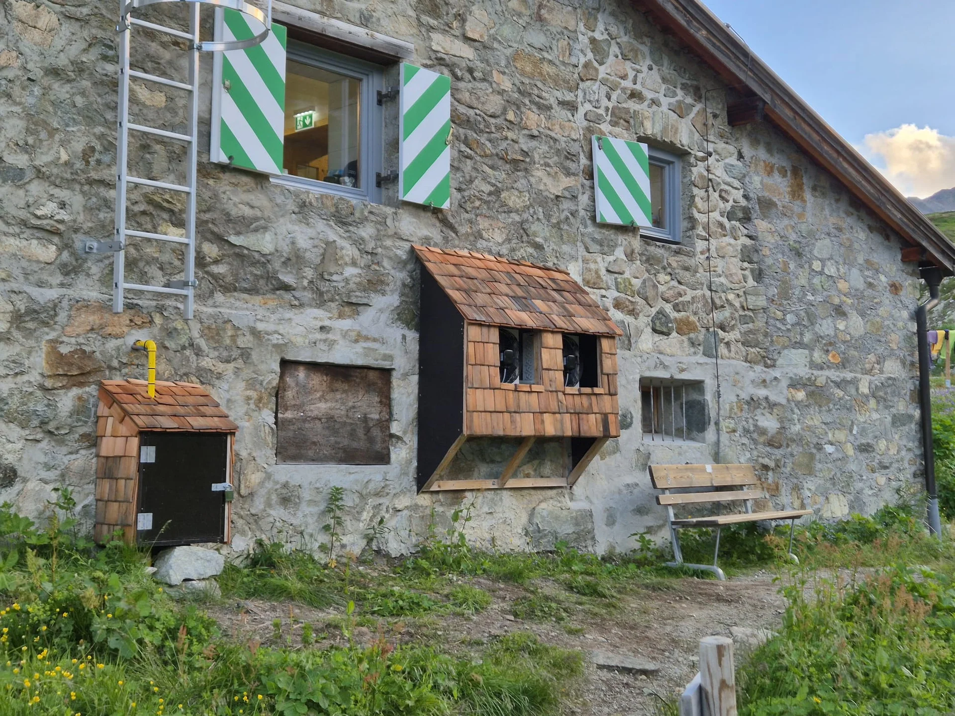 Außenwand der Häfler Hütte mit zwei Fenstern, eines mit grün-weiß gestreiften Fensterläden, darunter zwei kleine Holzkästen mit Schindelverkleidung und eine Holzbank | © DAV-FN / R. Klees o. R. Zahner