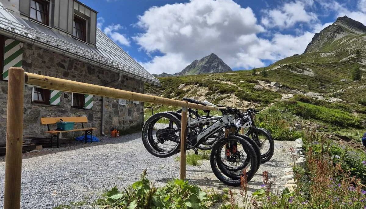 Fahrräder an einem Holzständer vor der Häfler Hütte mit steinigem Weg und grünen Bergen unter bewölktem Himmel | © DAV-FN / R. Klees o. R. Zahner