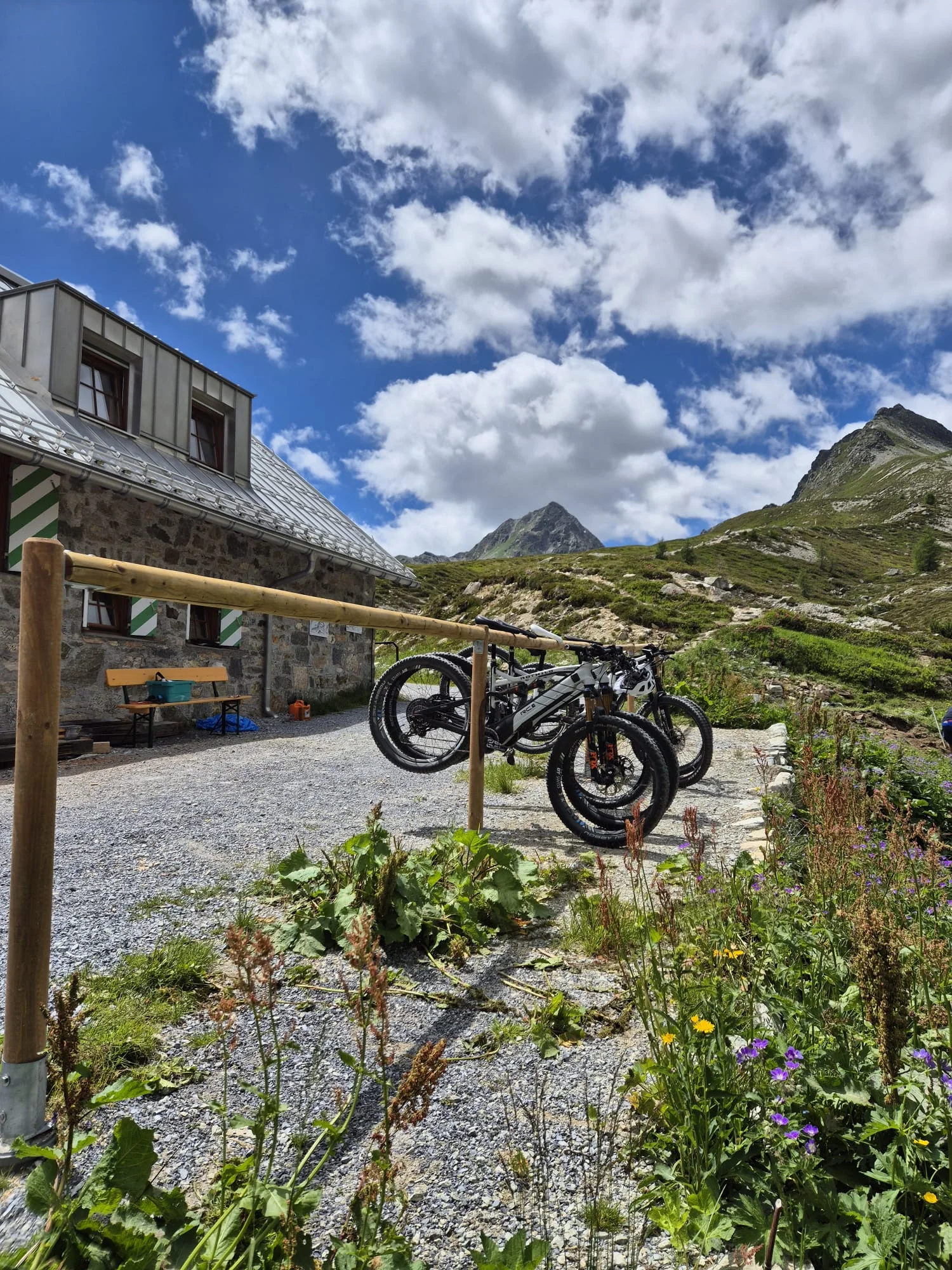 Fahrräder an einem Holzständer vor der Häfler Hütte mit steinigem Weg und grünen Bergen unter bewölktem Himmel | © DAV-FN / R. Klees o. R. Zahner
