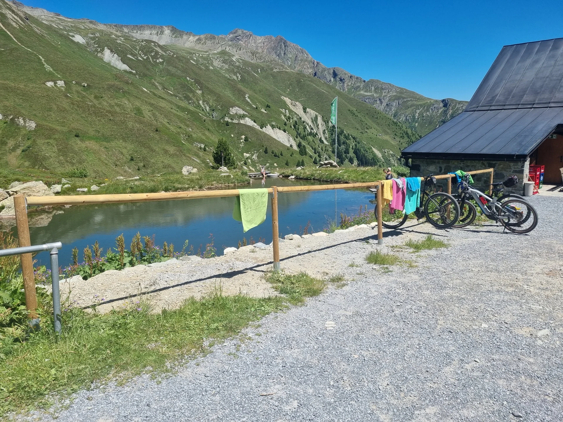 Bergsee mit grünen Hängen, Holzgeländer als MTB-Ständer mit bunten Handtüchern, zwei Mountainbikes neben der Häfler Hütte | © DAV-FN / R. Klees o. R. Zahner