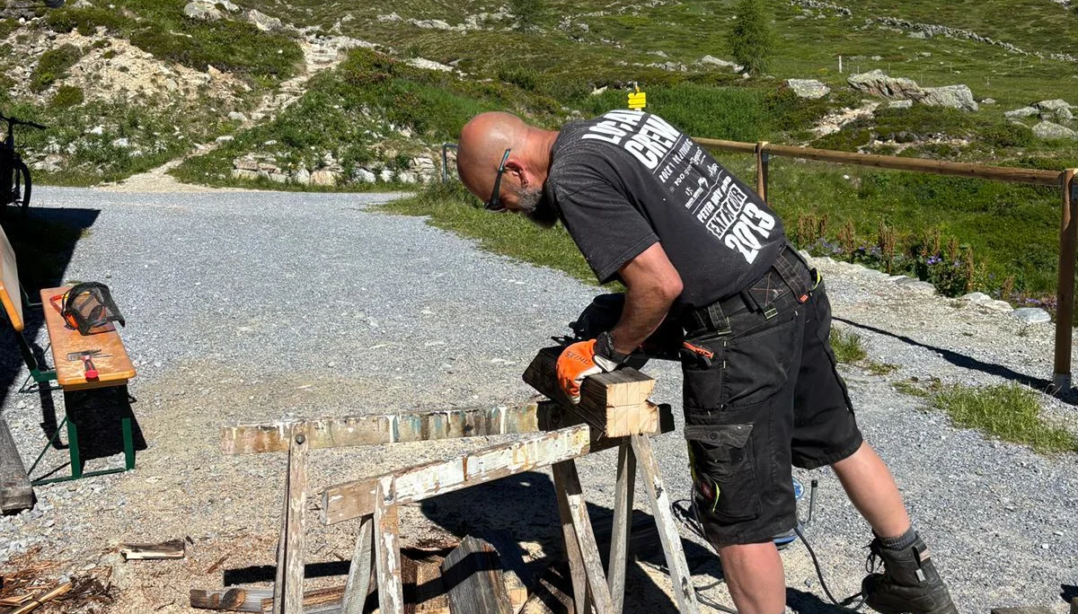 Mann sägt Brennholz auf einer Sägevorrichtung im Freien, umgeben von Bergen und grünem Gelände | © DAV-FN / R. Klees o. R. Zahner