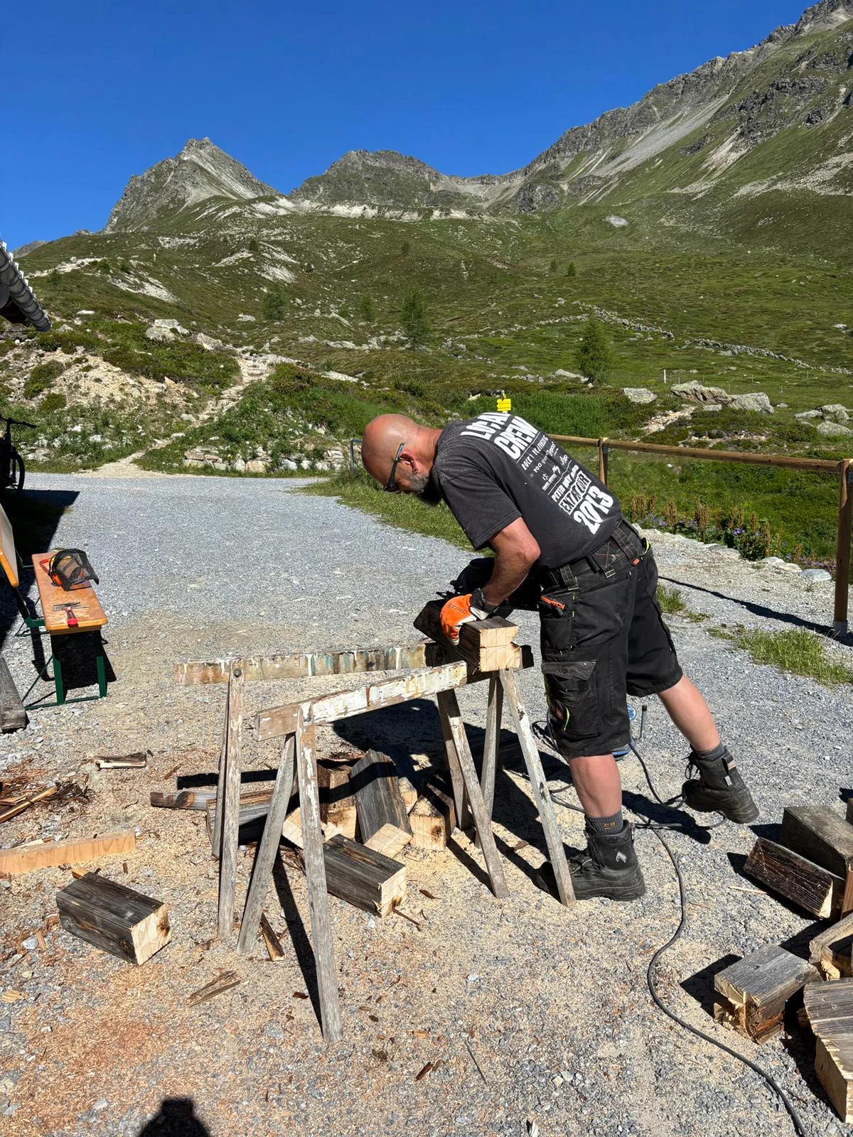 Mann sägt Brennholz auf einer Sägevorrichtung im Freien, umgeben von Bergen und grünem Gelände | © DAV-FN / R. Klees o. R. Zahner