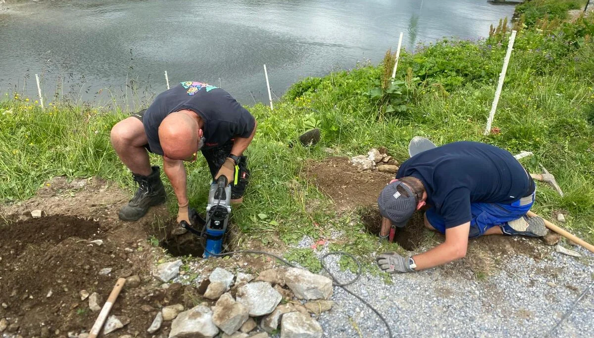 Zwei Männer arbeiten mit Werkzeugen an einem steinigen Boden neben einem See in einer Berglandschaft | © DAV-FN / R. Klees o. R. Zahner