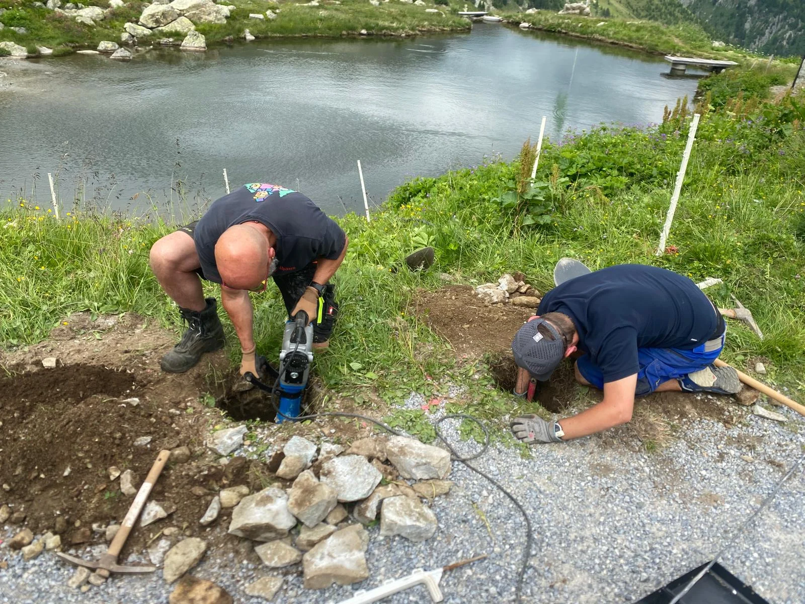 Zwei Männer arbeiten mit Werkzeugen an einem steinigen Boden neben einem See in einer Berglandschaft | © DAV-FN / R. Klees o. R. Zahner